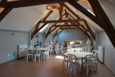 Salle lumineuse avec poutres apparentes, tables rondes dressées avec nappes blanches et chaises en plastique blanc.