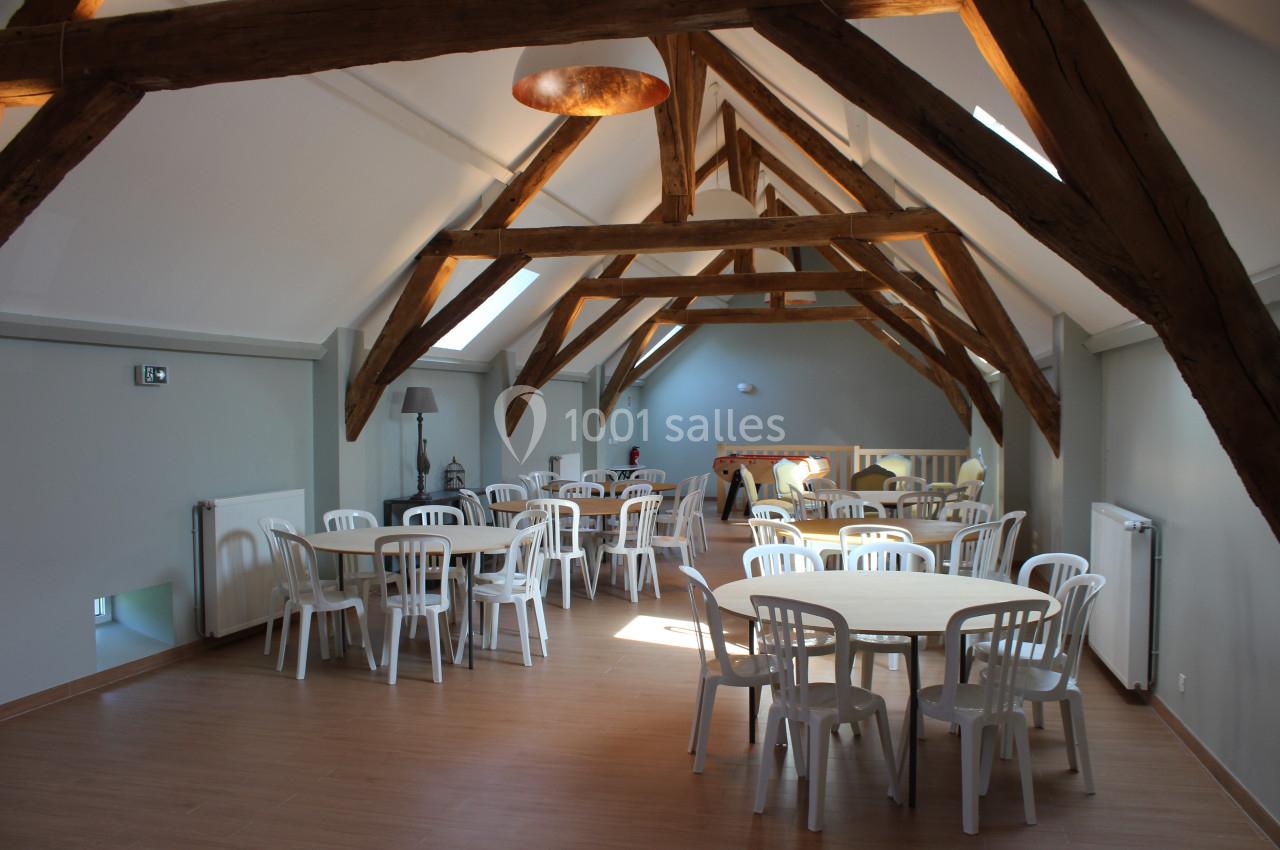 Salle lumineuse avec charpente en bois apparente, tables rondes blanches et chaises en plastique disposées uniformément.
