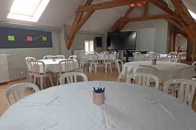Salle lumineuse avec poutres apparentes, tables rondes dressées avec nappes blanches et chaises en plastique blanc.