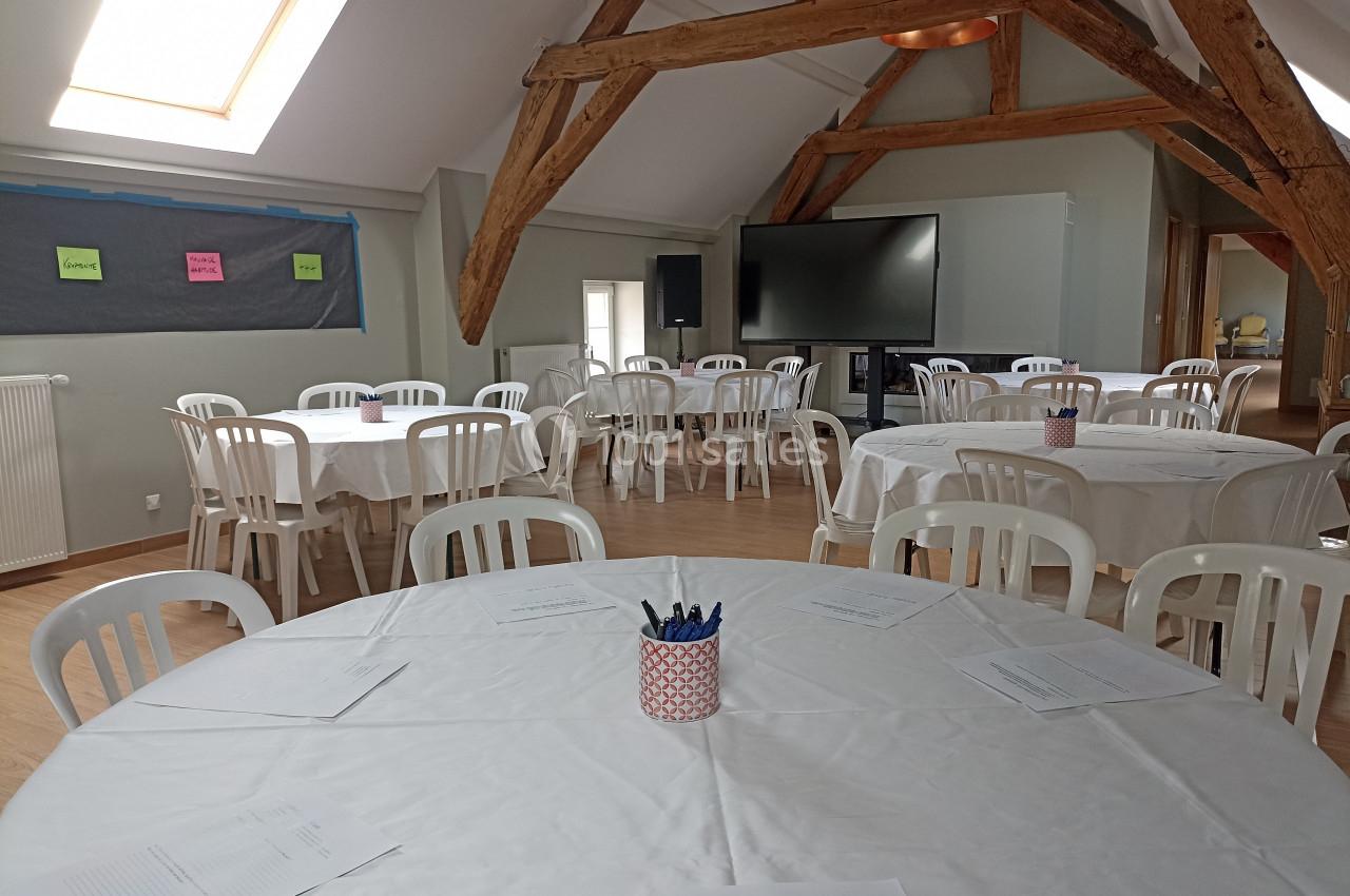 Salle lumineuse avec poutres apparentes, tables rondes couvertes de nappes blanches et chaises en plastique disposées pour…