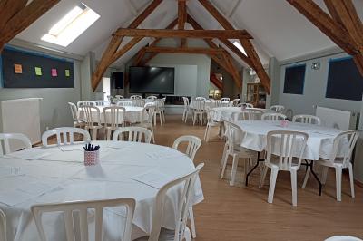 Salle lumineuse avec poutres apparentes, tables rondes dressées avec nappes blanches et chaises en plastique blanc.