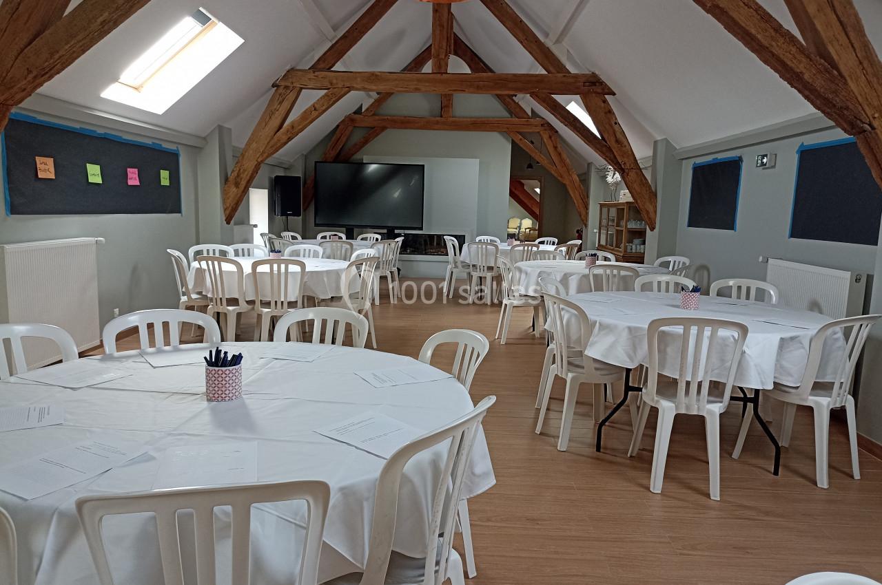 Salle lumineuse avec poutres apparentes, tables rondes recouvertes de nappes blanches et chaises en plastique blanc.