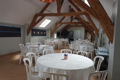 Salle lumineuse avec poutres apparentes, tables rondes dressées avec nappes blanches et chaises en plastique blanc.