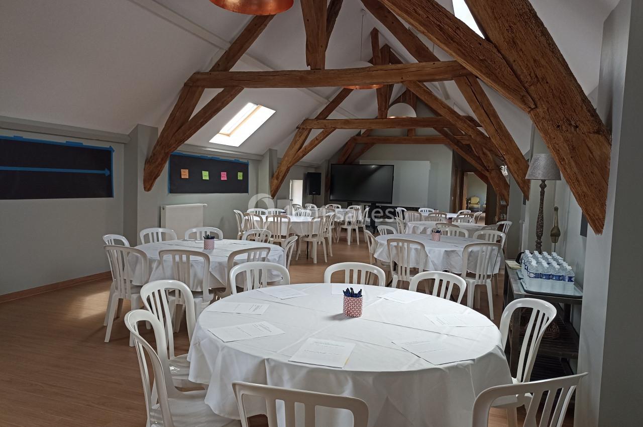 Salle lumineuse avec poutres apparentes, tables rondes dressées avec nappes blanches et chaises en plastique blanc.