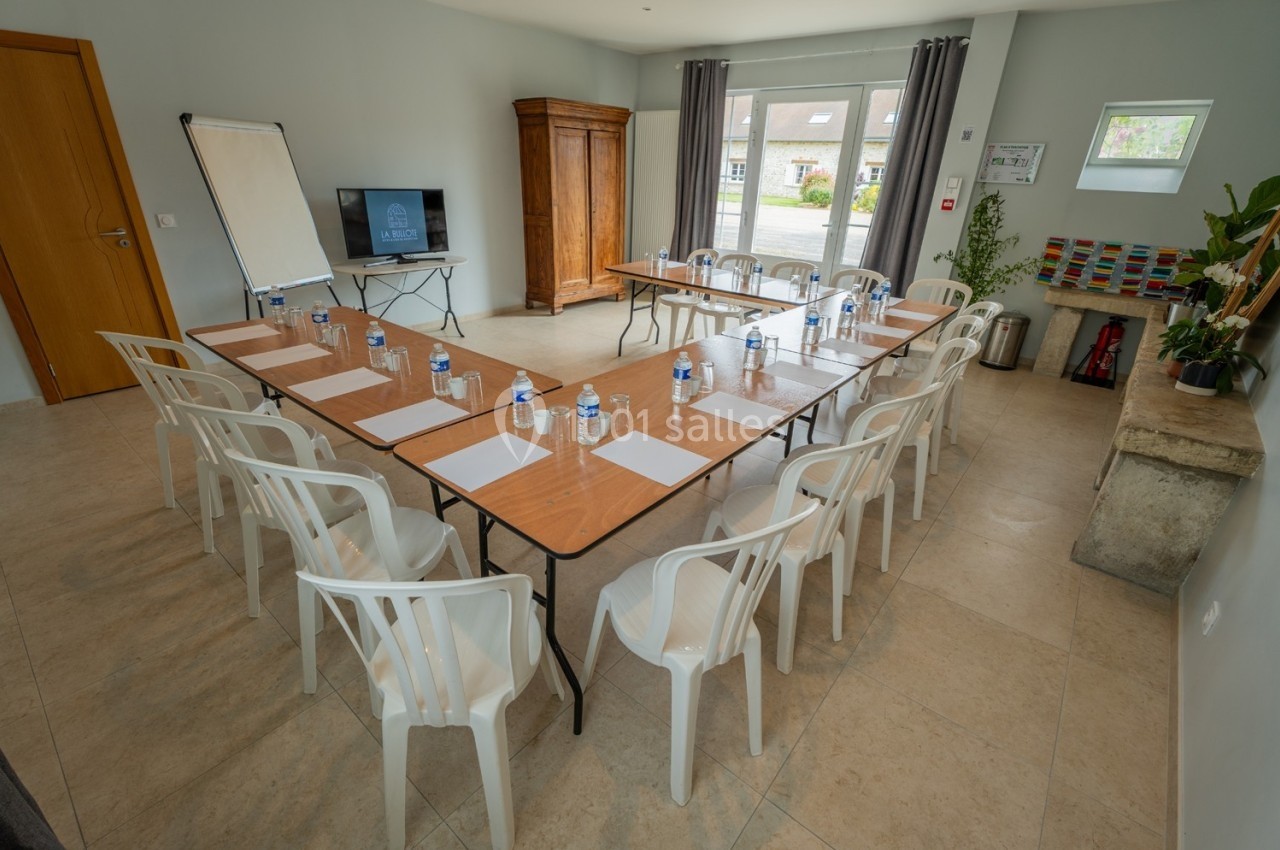 Salle de réunion lumineuse avec tables disposées en U, chaises en plastique, bouteilles d'eau et écran de présentation.