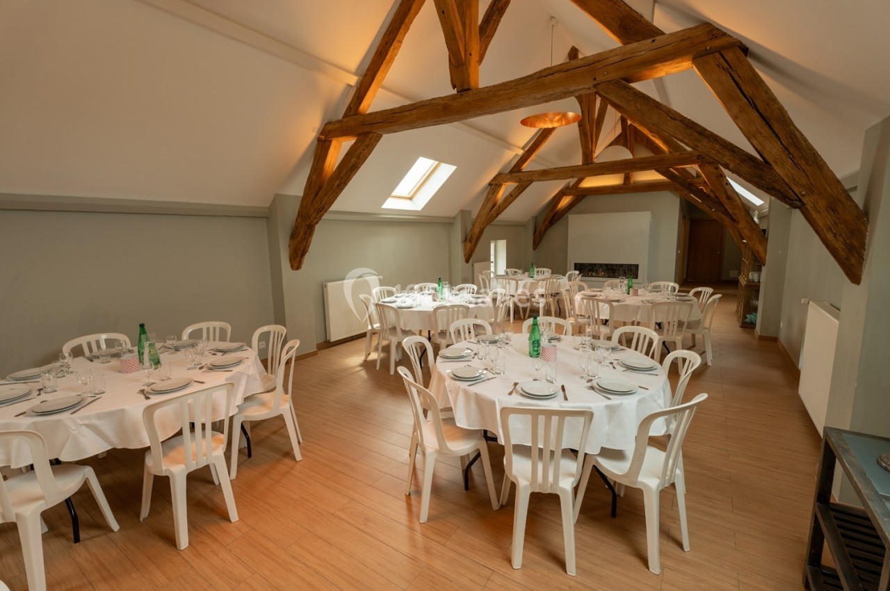 Salle de réception avec poutres apparentes, tables rondes dressées avec nappes blanches et chaises en plastique.