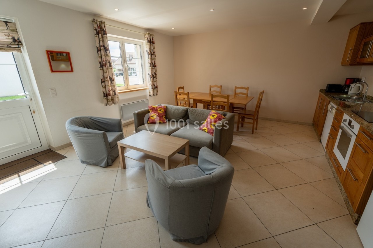 Salon avec canapé, fauteuils gris, table basse, table à manger en bois et cuisine équipée dans une pièce lumineuse.