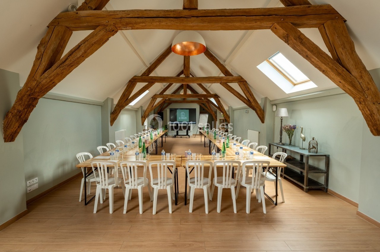 Salle de réunion lumineuse avec poutres apparentes, tables en U, chaises blanches et équipement de présentation.