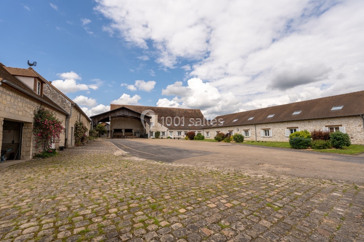 Cour pavée entourée de bâtiments en pierre avec toits en tuiles, sous un ciel partiellement nuageux.