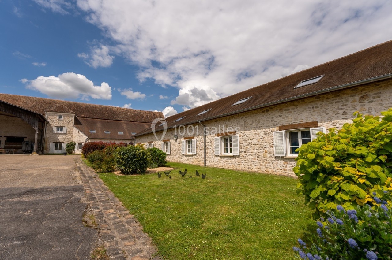 Bâtiment en pierre avec toit en tuiles, entouré de pelouse, arbustes et ciel partiellement nuageux.