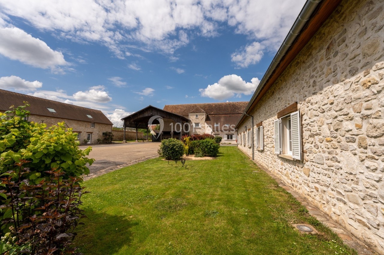 Cour d'une ferme avec bâtiments en pierre, pelouse bien entretenue et ciel partiellement nuageux.
