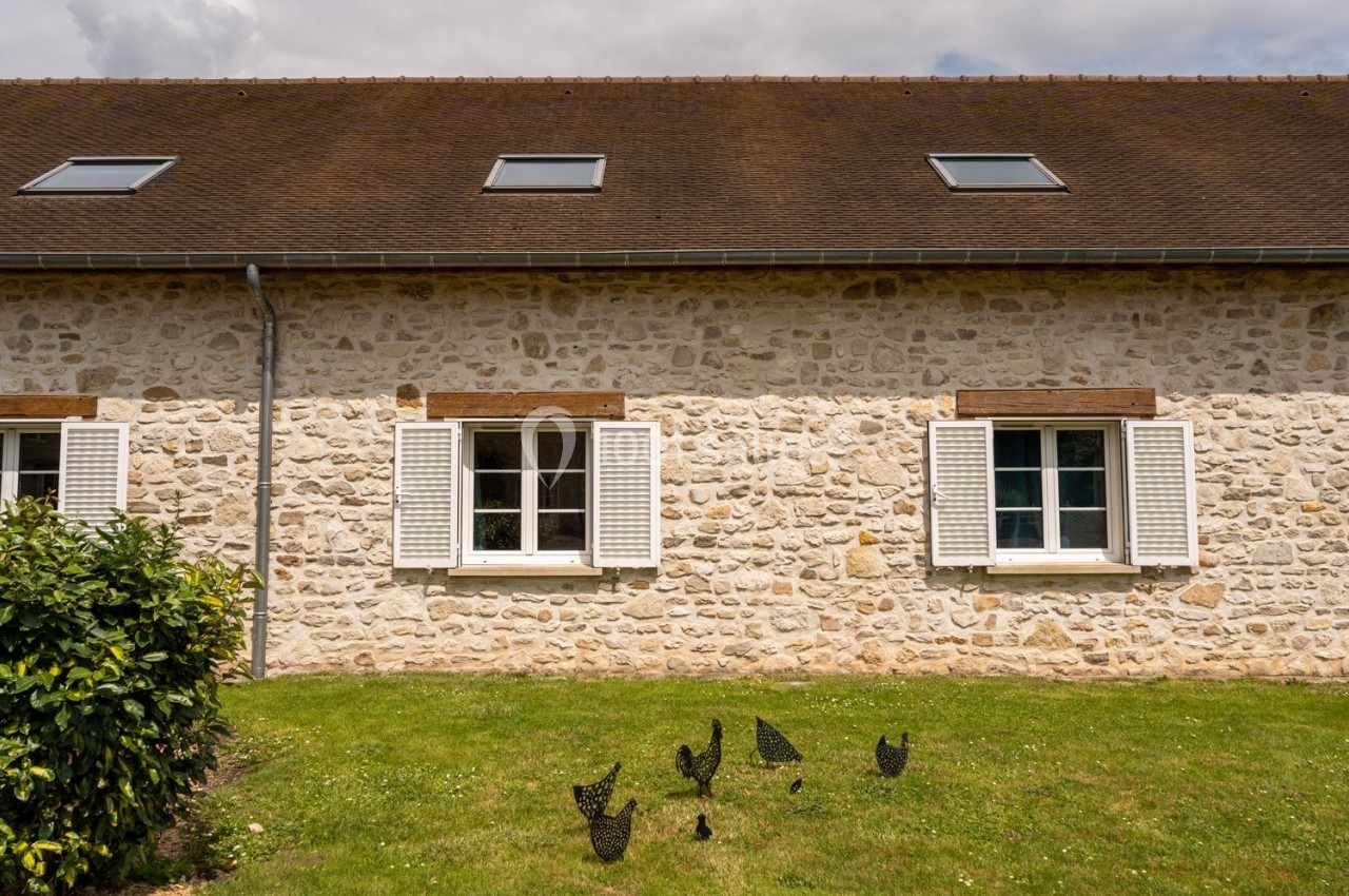 Façade en pierre d'une maison avec volets blancs, fenêtres mansardées et pelouse ornée de sculptures métalliques.