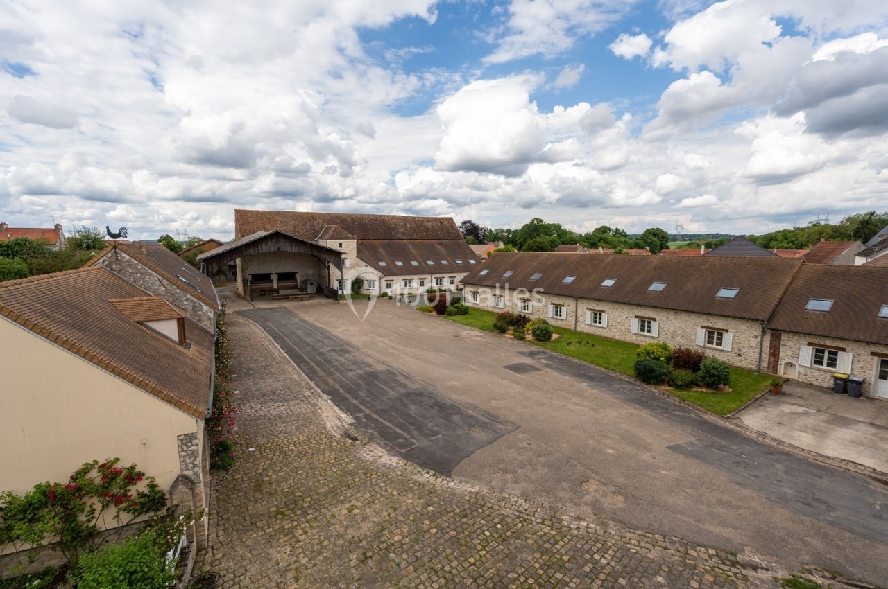 Cour pavée entourée de bâtiments en pierre avec toits en tuiles, sous un ciel partiellement nuageux.
