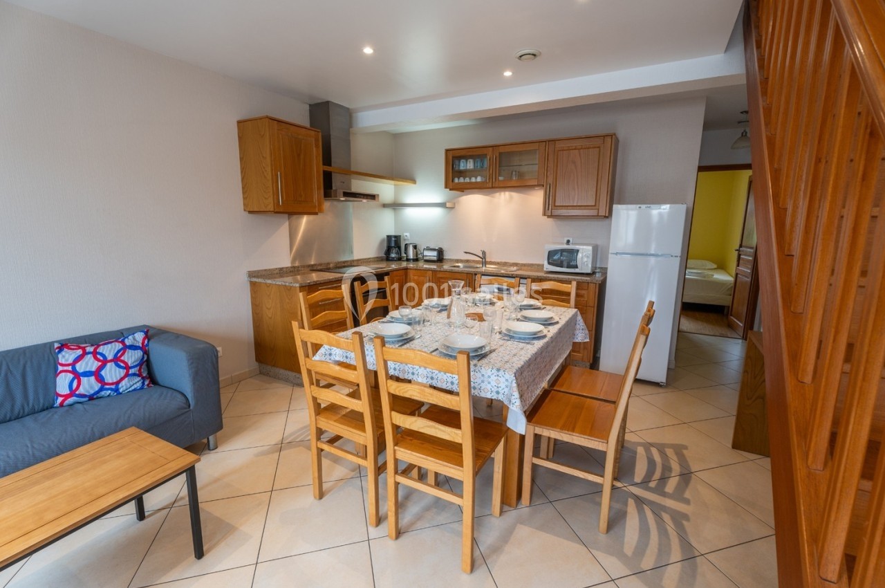 Cuisine ouverte avec table dressée pour six personnes, meubles en bois, canapé et escalier visibles en arrière-plan.