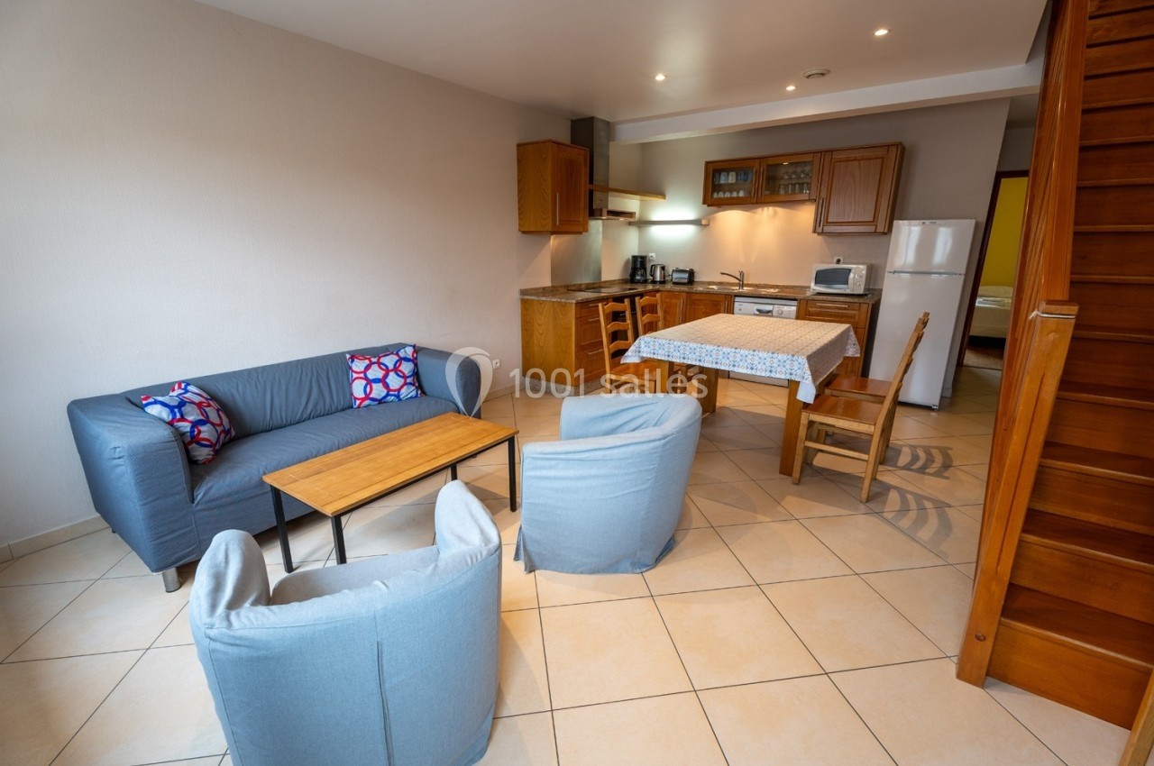 Salon avec canapé, fauteuils, table basse et cuisine ouverte équipée, dans un intérieur lumineux avec carrelage beige.