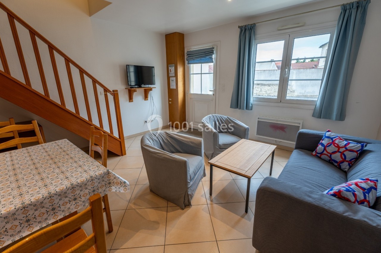 Salon lumineux avec escalier en bois, canapé, fauteuils, table basse, table à manger et fenêtre donnant sur l'extérieur.