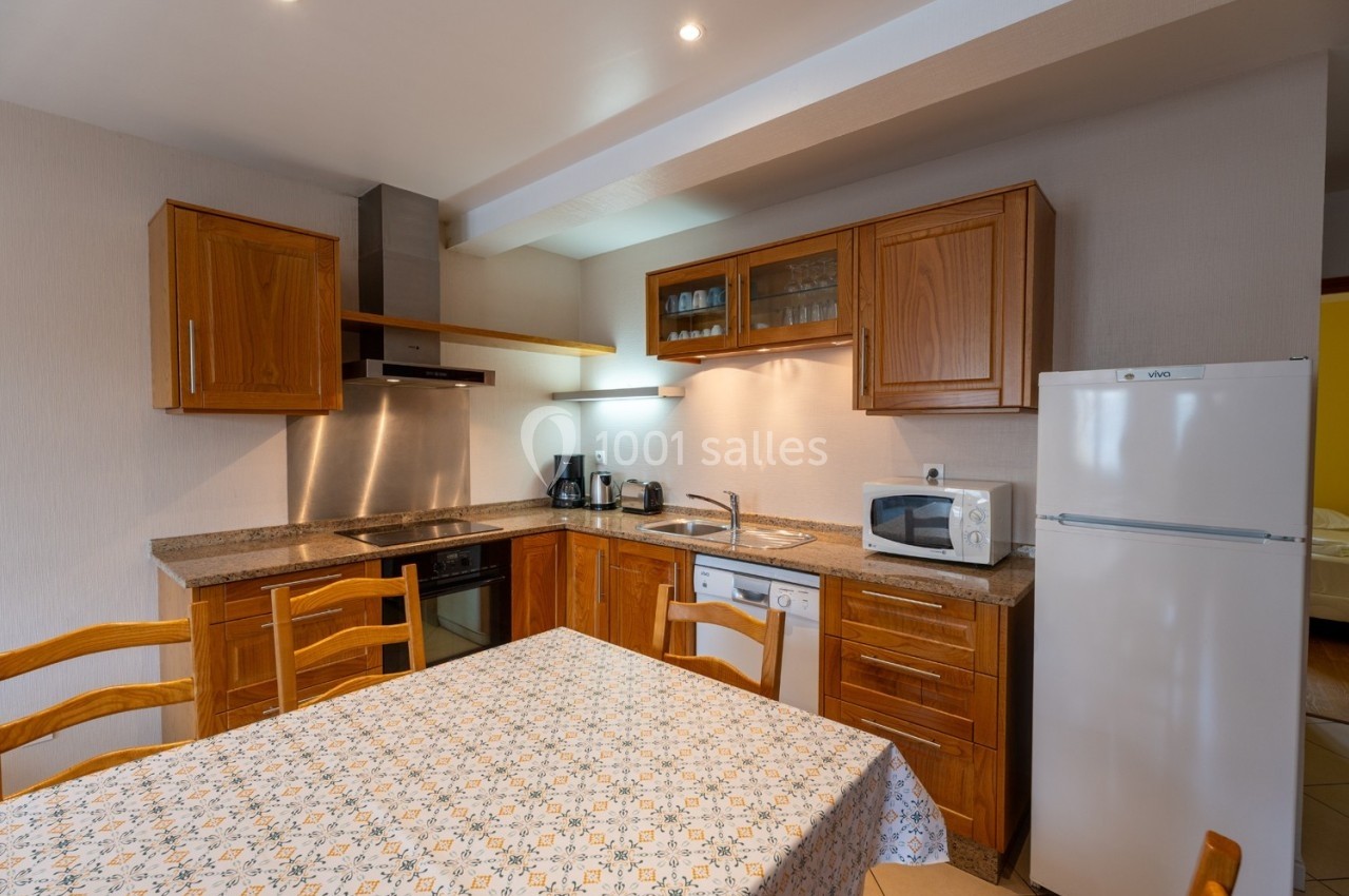 Cuisine équipée avec meubles en bois, table recouverte d'une nappe, réfrigérateur, micro-ondes et électroménagers.