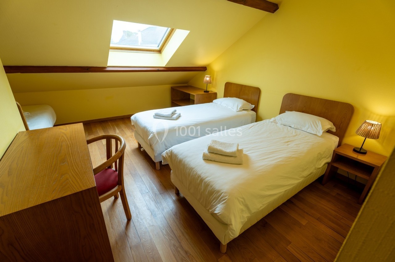 Deux lits simples avec linge plié dans une chambre mansardée lumineuse, murs jaunes et parquet en bois.