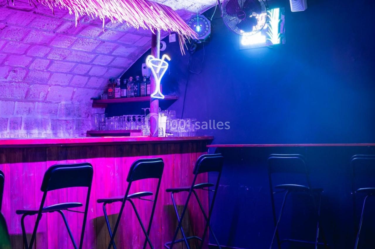 Bar avec tabourets noirs, éclairage violet, néon en forme de verre et étagères avec bouteilles sous une voûte en pierre.