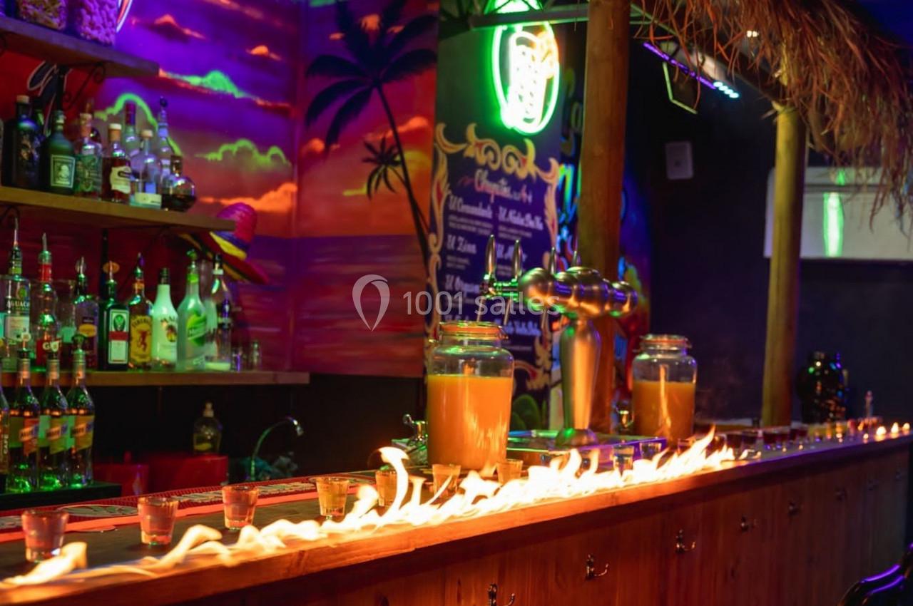 Bar tropical avec des flammes sur le comptoir, décor coloré et bouteilles alignées en arrière-plan.