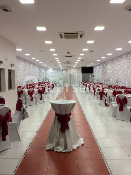 Salle de réception décorée avec des chaises blanches ornées de nœuds rouges et une table haute au centre.