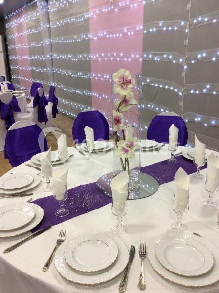 Table dressée avec une nappe blanche, serviettes pliées, chemin de table violet, et centre floral avec des orchidées roses.