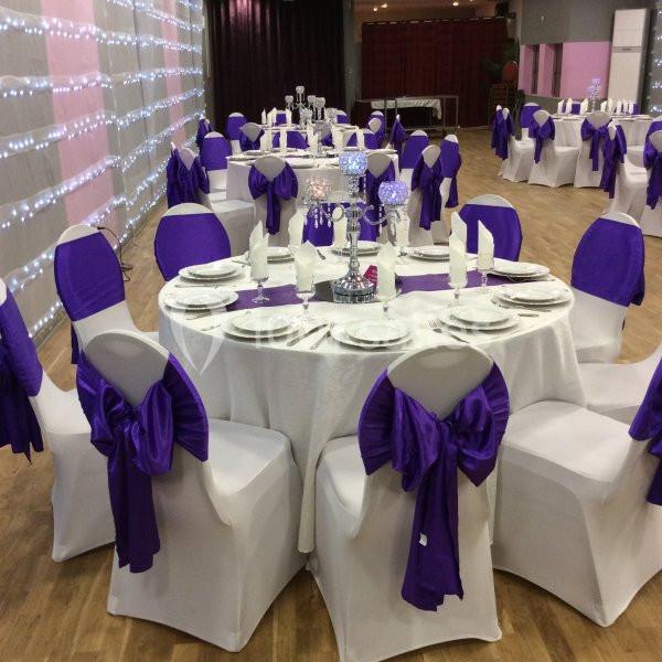 Salle de réception décorée avec des nappes blanches, des chaises ornées de nœuds violets et des tables dressées élégamment.