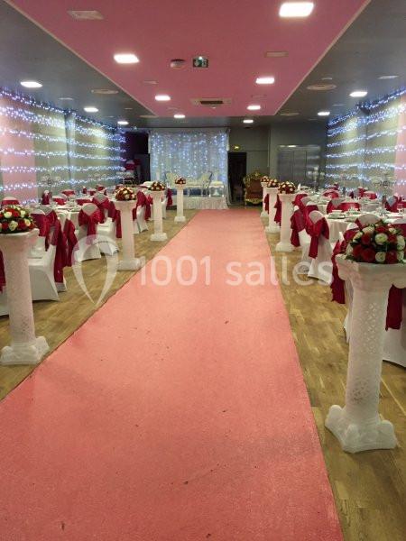 Salle de réception décorée avec des tables nappées de blanc et rouge, allée centrale rose et guirlandes lumineuses.