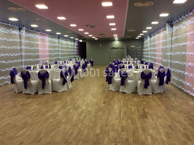 Salle de réception décorée avec des tables rondes, nappes blanches et nœuds violets, éclairée par des guirlandes lumineuses.