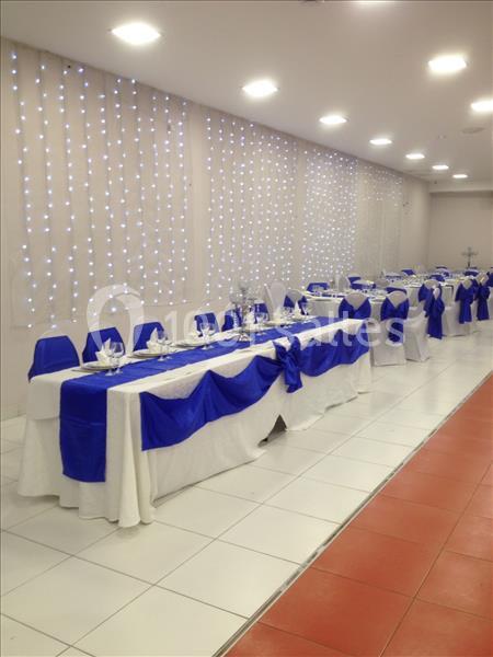 Salle de réception avec tables dressées, nappes blanches et chemins de table bleus, éclairée par des guirlandes lumineuses.