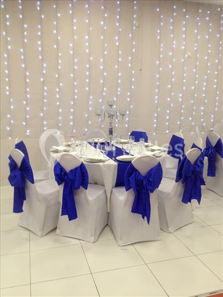 Table dressée avec nappes blanches, chaises décorées de nœuds bleus et fond illuminé par des guirlandes lumineuses.
