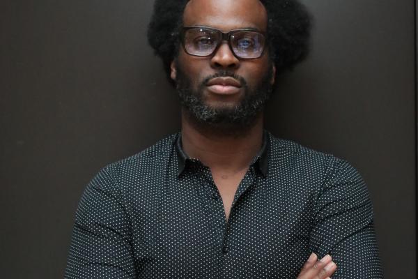 Homme portant des lunettes et une chemise à motifs, bras croisés, debout devant un fond sombre.