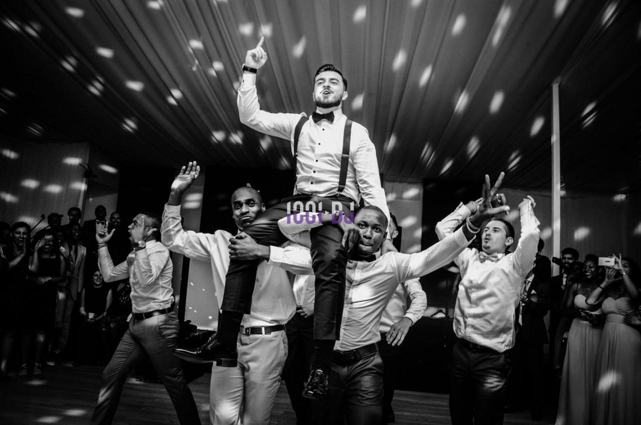 Un homme en tenue formelle est porté par quatre autres hommes dans une ambiance festive sous des lumières tamisées.