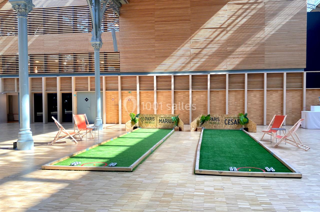 Deux terrains de pétanque en intérieur avec chaises longues et décorations végétales sur un sol en bois.