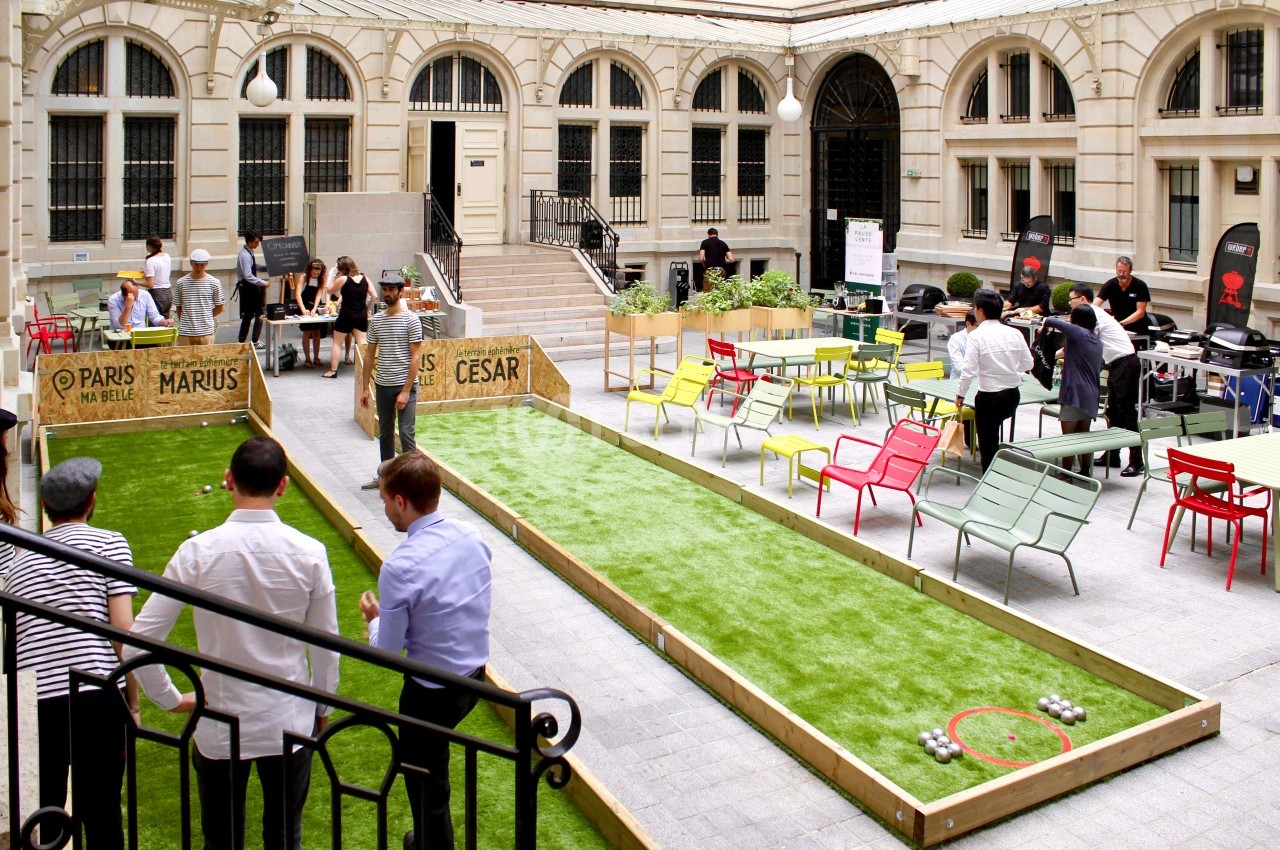 Cour intérieure aménagée avec terrains de pétanque, tables colorées et personnes jouant ou discutant.