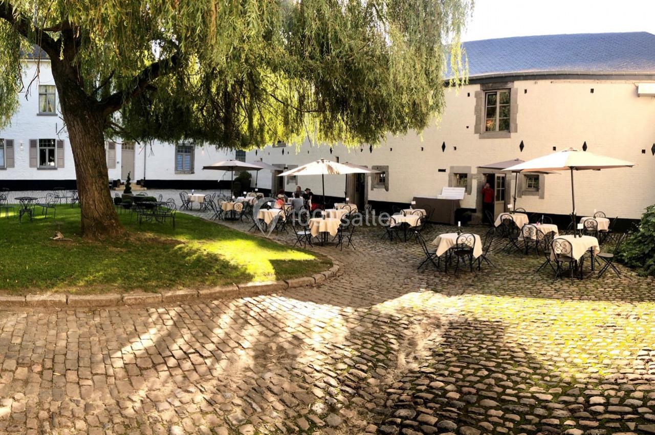 Cour pavée avec des tables et chaises en fer forgé, partiellement ombragées par des parasols et un grand arbre.