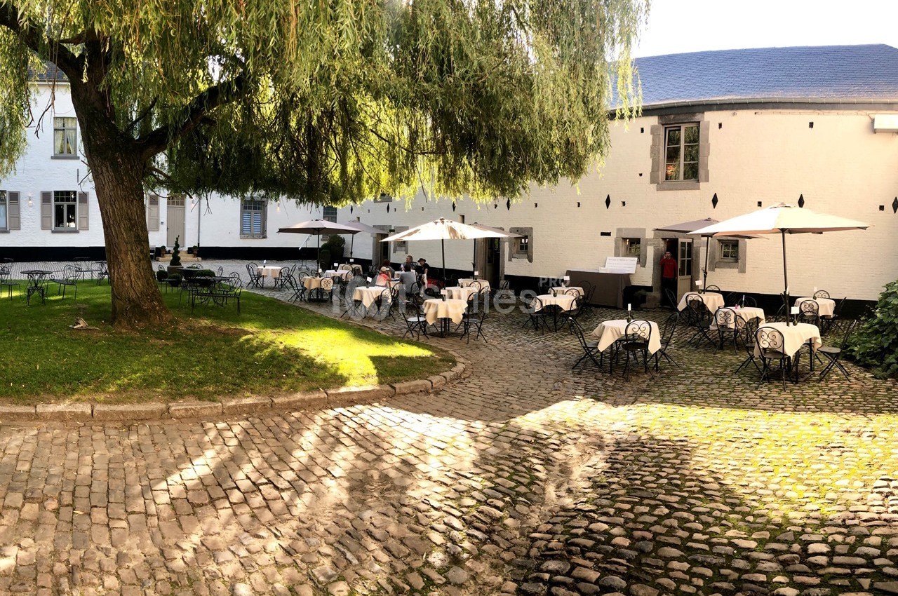 Cour pavée avec des tables et chaises en fer forgé, partiellement ombragées par des parasols et un grand arbre.