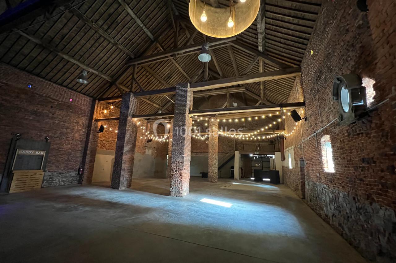 Intérieur d'une grande salle en briques avec charpente en bois, éclairée par des guirlandes lumineuses suspendues.
