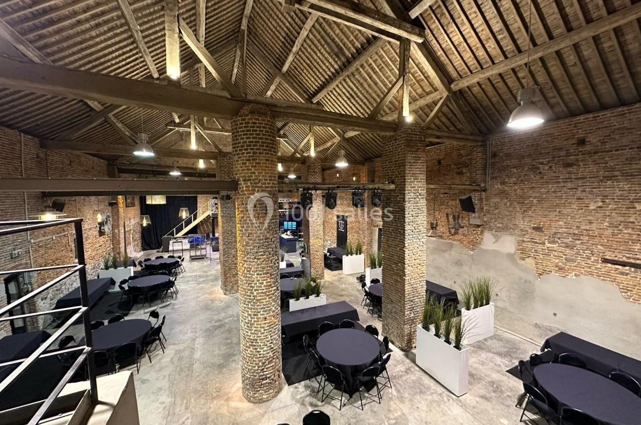 Salle spacieuse avec murs en briques, plafond en poutres apparentes et tables rondes noires disposées pour un événement.