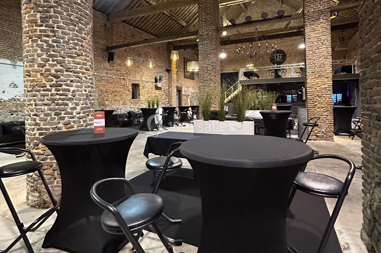 Salle en briques avec tables hautes noires et chaises, décorée de plantes et éclairée par des lustres suspendus.