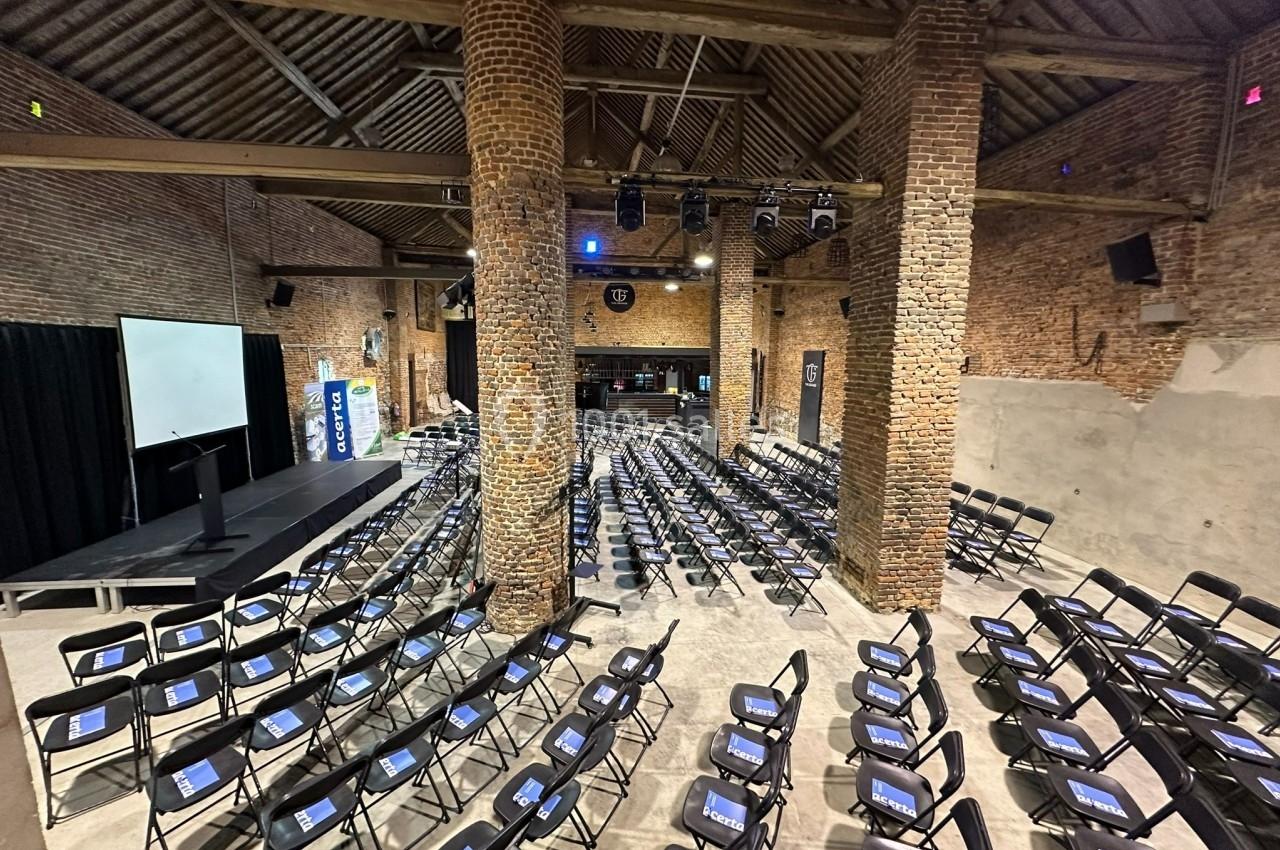 Salle en briques avec des rangées de chaises noires alignées, écran de projection et estrade à l'avant.