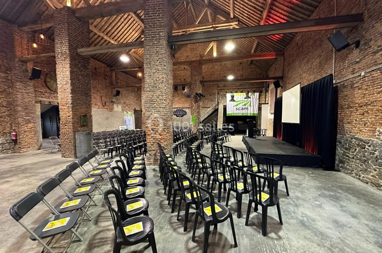 Salle en briques avec des rangées de chaises noires alignées face à une scène et un écran de projection.