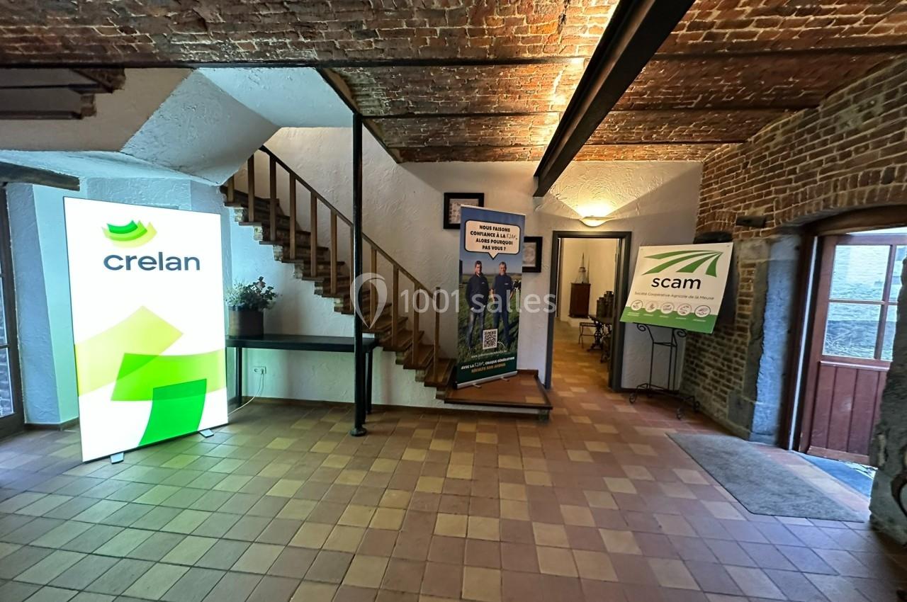 Entrée d'un bâtiment avec des affiches de Crelan et Scam, un escalier en bois et un sol en carreaux.