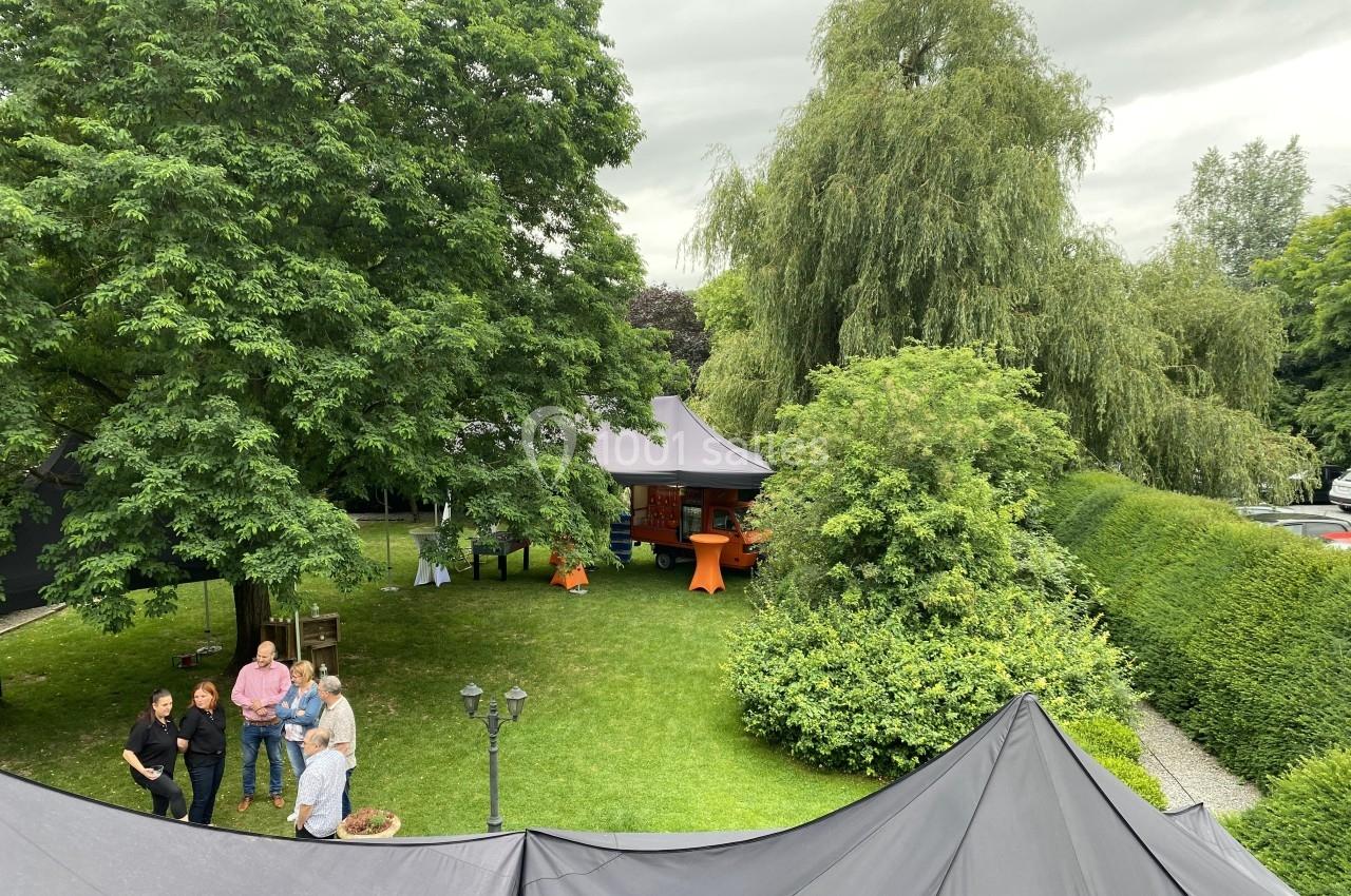 Vue d'un jardin verdoyant avec des arbres, des tentes et un petit groupe de personnes discutant près d'une allée.