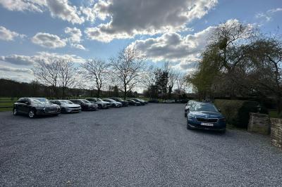 Parking en gravier entouré d'arbres et de bâtiments en briques sous un ciel partiellement nuageux.