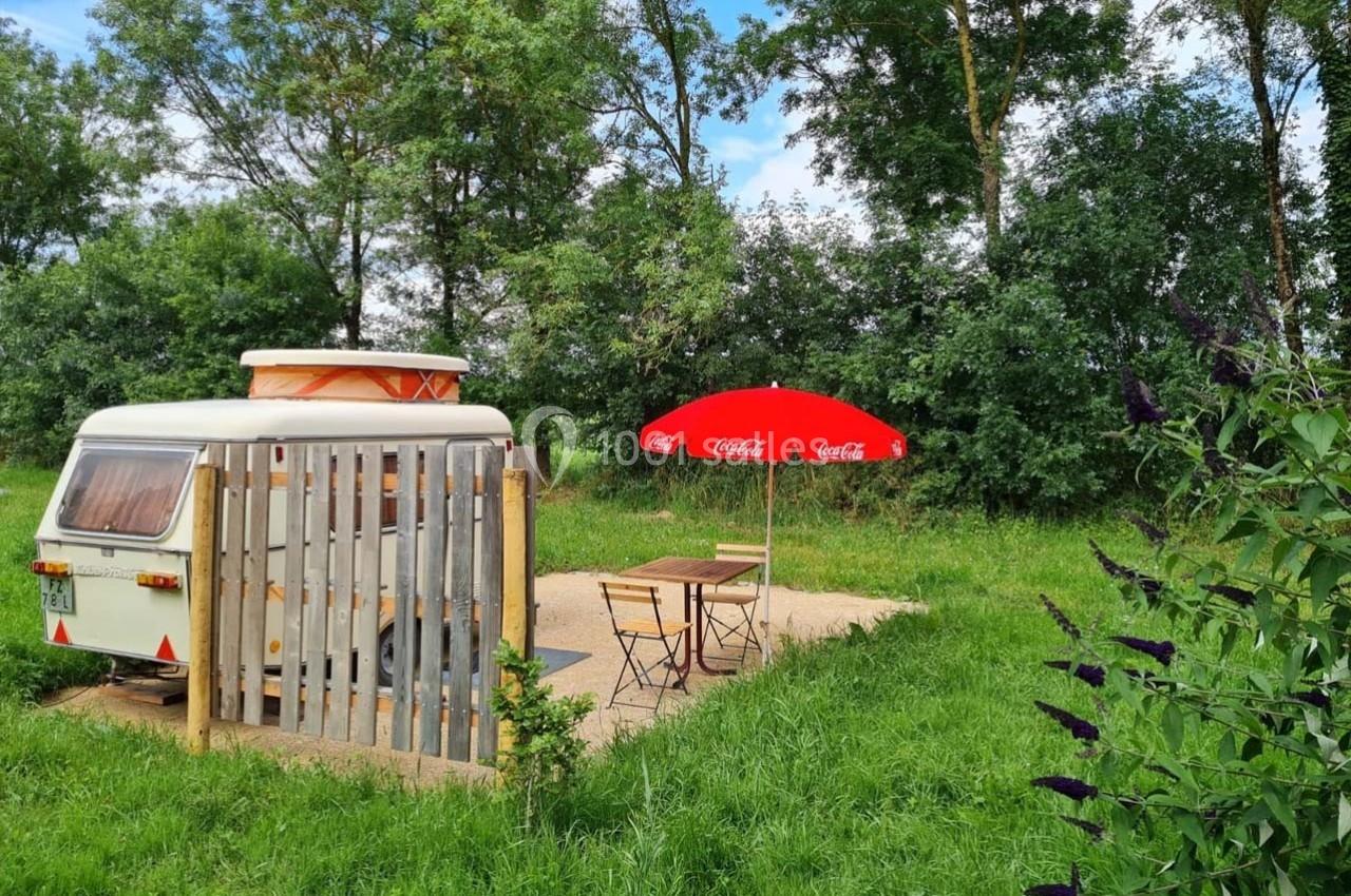 Caravane blanche stationnée près d'une terrasse avec parasol rouge et mobilier de jardin, entourée de verdure.
