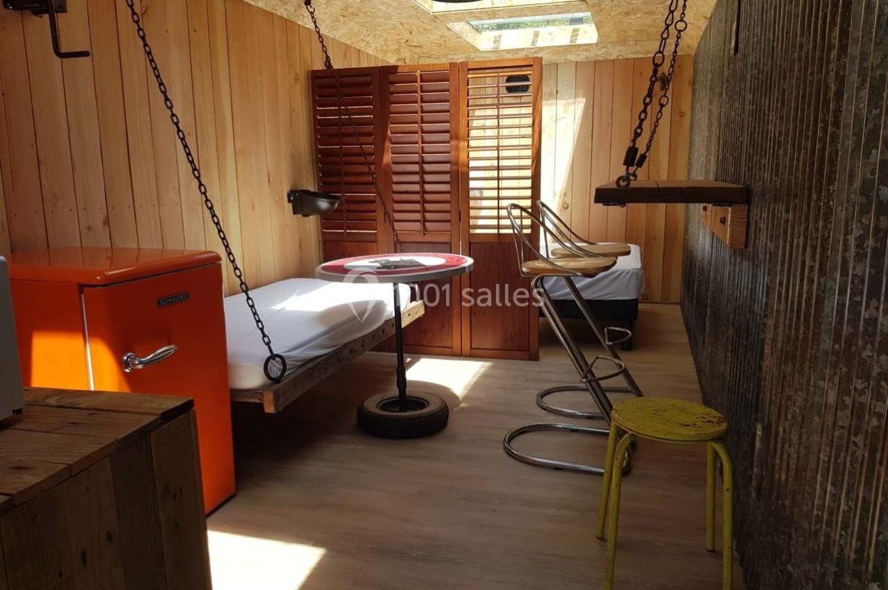 Chambre en bois avec deux lits suspendus, un réfrigérateur orange, des tabourets métalliques et une lumière naturelle.