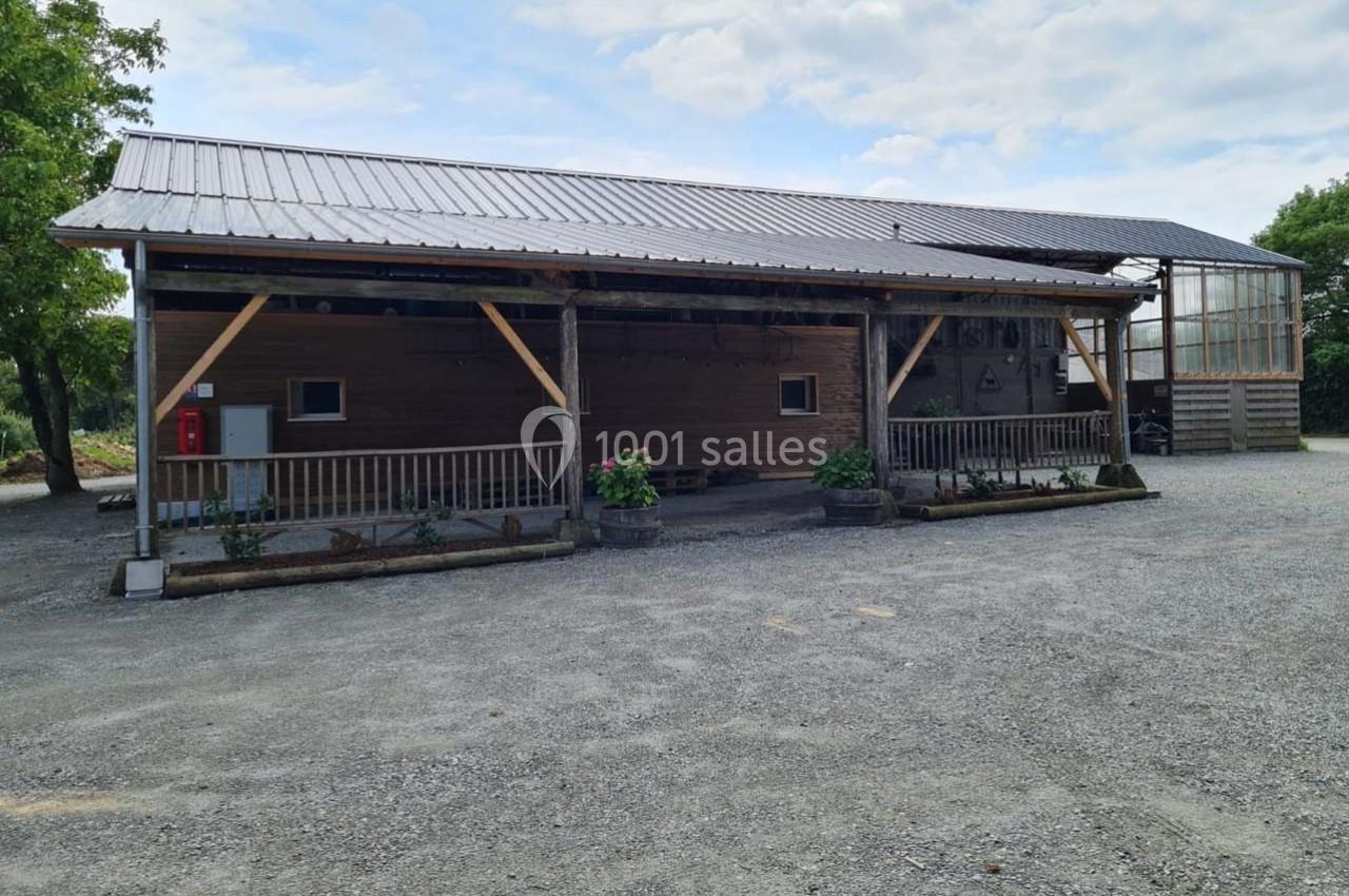 Bâtiment en bois avec toit métallique, entouré de gravier et de quelques plantes en pots, sous un ciel partiellement nuageux.