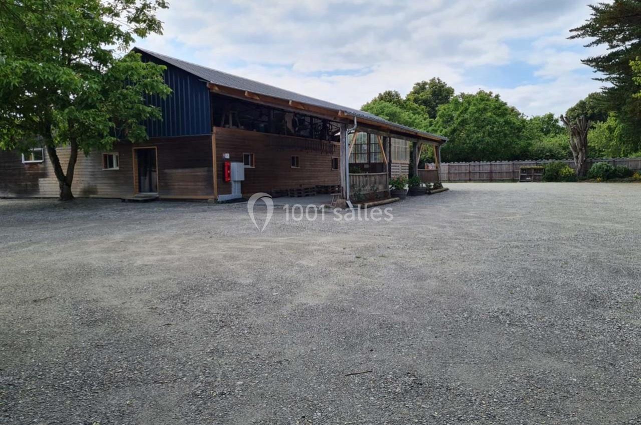 Bâtiment en bois avec toit métallique, entouré d'arbres, donnant sur une grande cour en gravier sous un ciel partiellement…