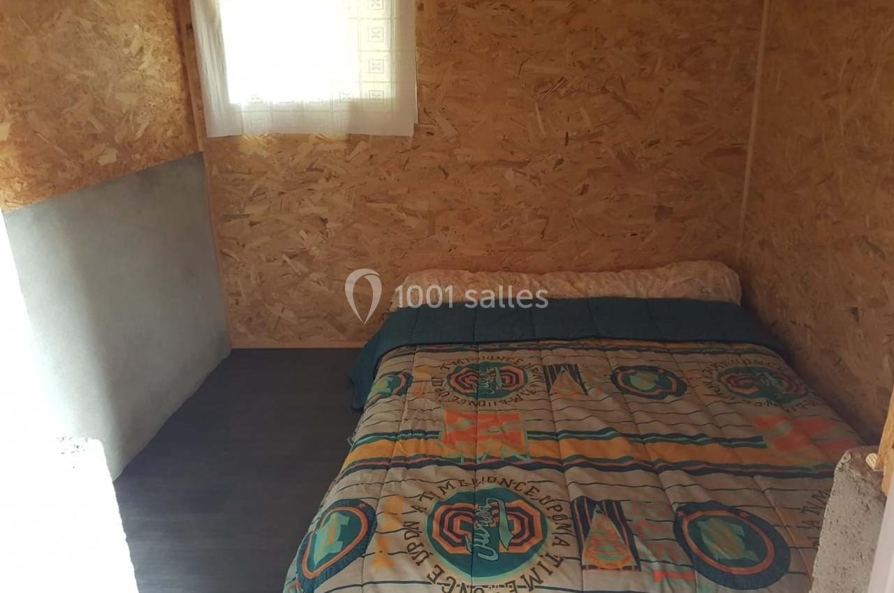 Lit double avec une couverture colorée dans une petite pièce aux murs en bois aggloméré, éclairée par une fenêtre.